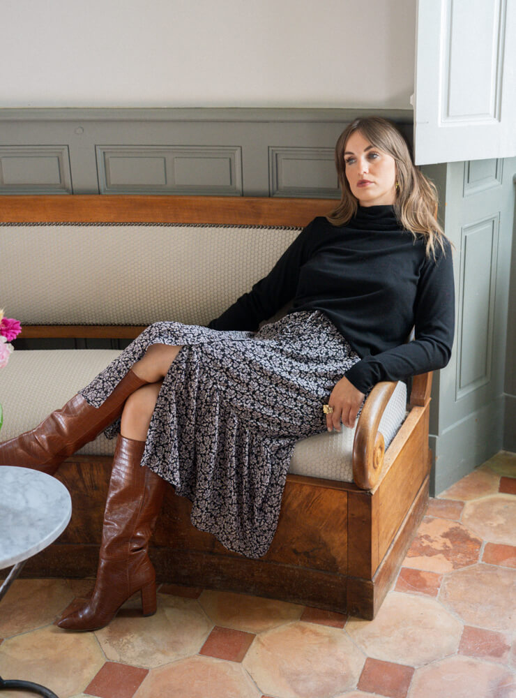 Modèle dans un canapé en bois qui porte un pull noir, une jupe à fleurs et des bijoux June&River