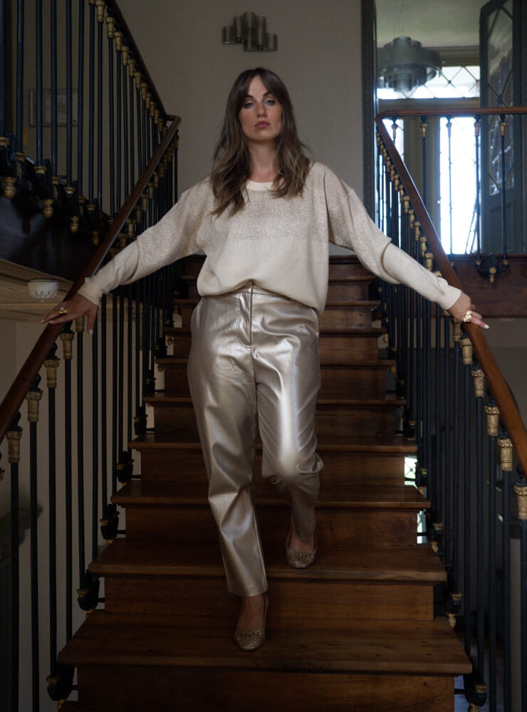 Modèle dans un escalier en bois qui porte un pull écru, un pantalon et des bijoux June&River