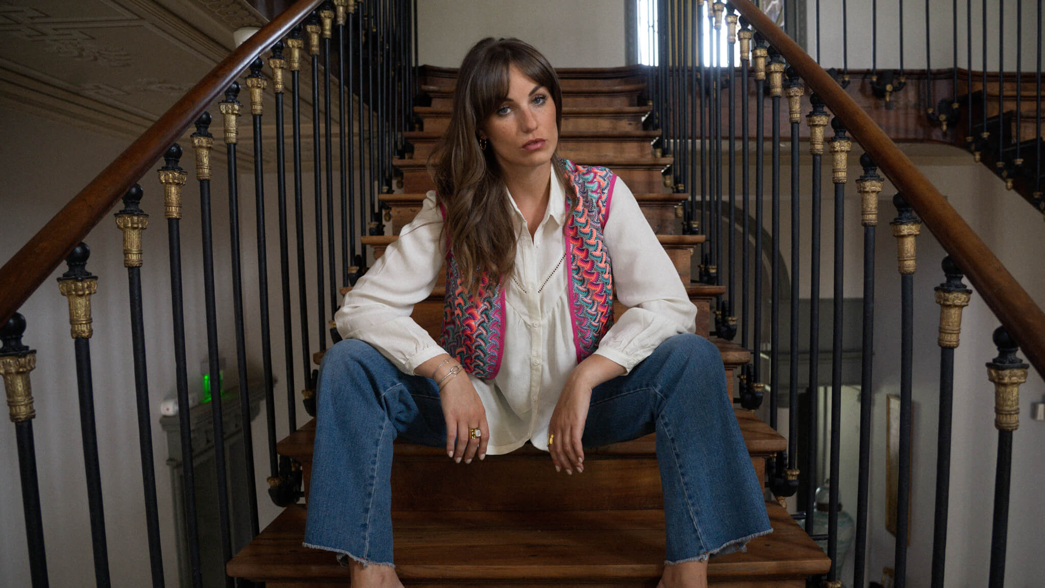 Modèle dans un escalier en bois qui porte un gilet coloré, une chemise écru et un jean June&River